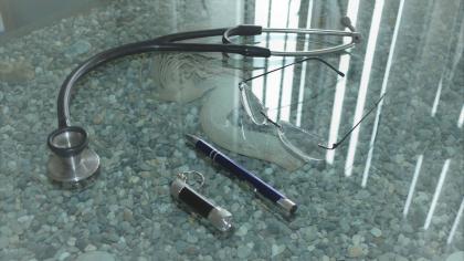 Stethoskop, Brille, Kugelschreiber und medizinische Taschenlampe liegen auf einem Glastisch mit Steinmuster.
