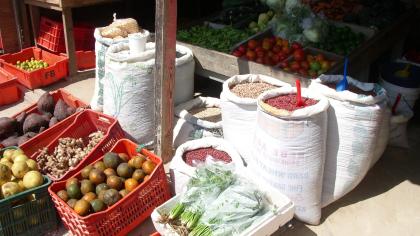 Bunter Marktstand mit Säcken voller Hülsenfrüchte und Getreide sowie Körben mit frischem Obst, Gemüse und Kräutern.