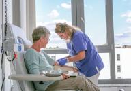 Pflegekraft in blauer Kleidung legt einer älteren Patientin in einem Behandlungsstuhl eine Infusion an, vor einem großen Fenster mit Blick auf den Himmel.