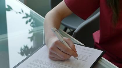Person in medizinischer Kleidung füllt ein Formular auf einem Klemmbrett mit einem Stift aus, sitzend an einem Glastisch.