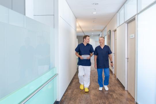 Zwei Ärzte in blauer Berufskleidung gehen lächelnd durch einen hellen Praxisflur und unterhalten sich